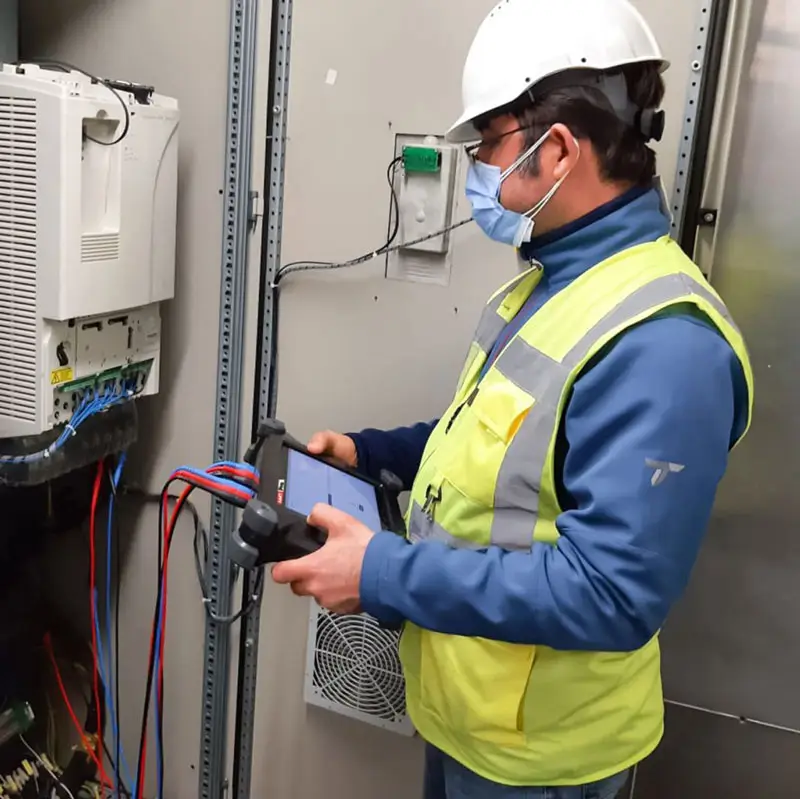 Worker in hard hat and vest inspects electrical panel with AMT Pro device.