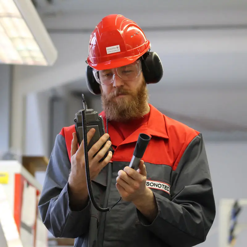 Bearded man in red hard hat inspects Sonotec Sonaphone®III device and cylinder.