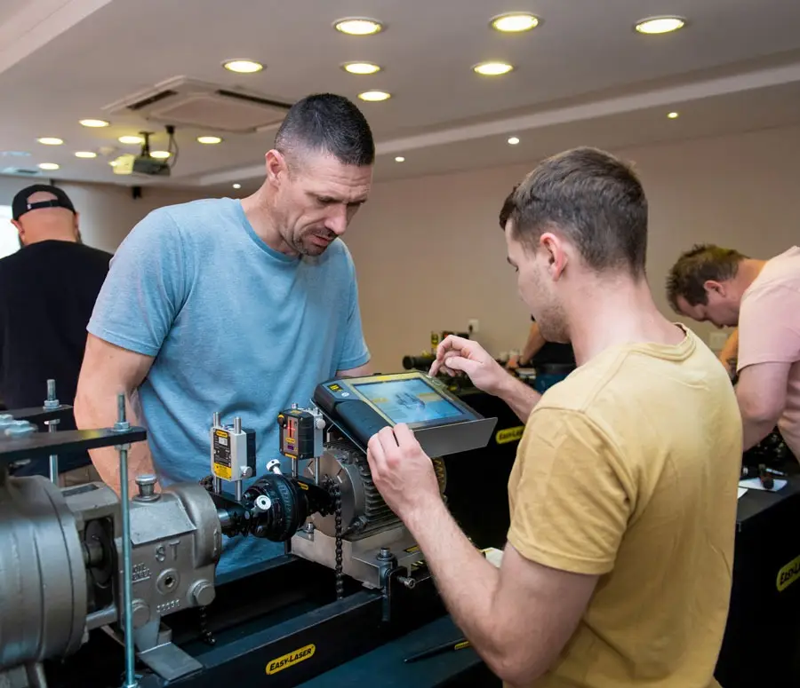 Two men operate industrial equipment, one using a touchscreen in a training room.