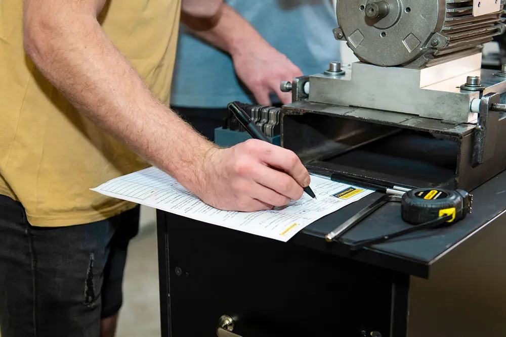 "Person in yellow shirt writing measurements on paper at workbench with tape measure and machining tool"