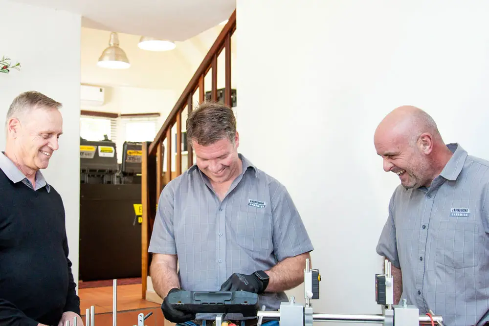 "Two technicians in gray work shirts working on equipment at indoor workstation near staircase"