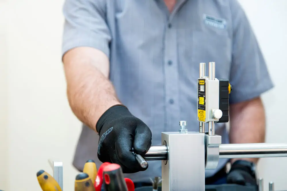 "Technician in gray shirt and gloves adjusting cylindrical component with measuring device attached in workshop"