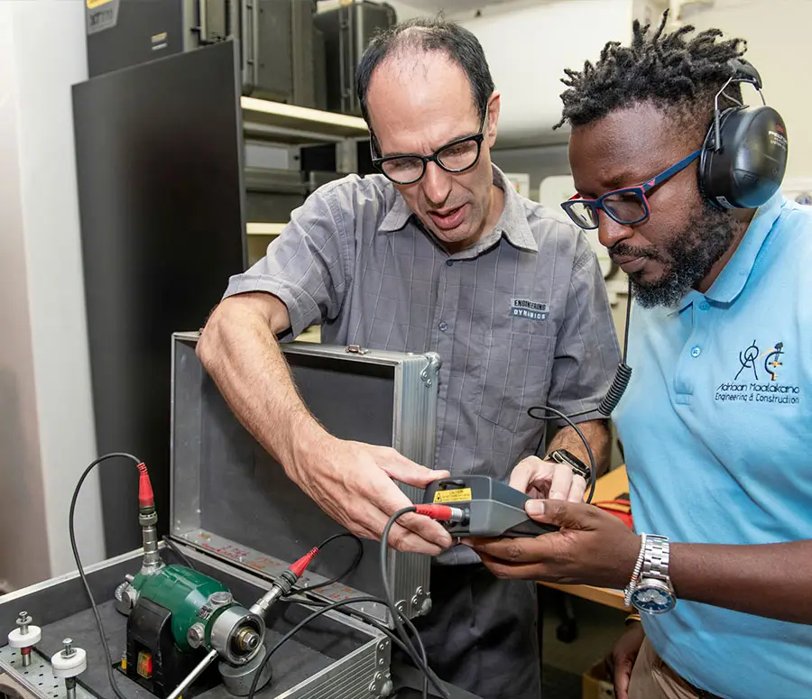 "Two technicians examining electronic device with probe and green cylindrical component in laboratory workspace"